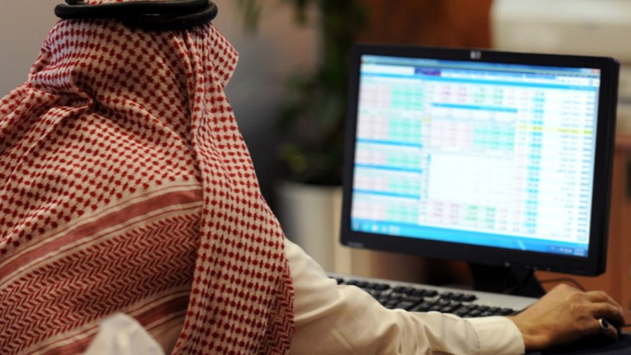 رجل بالزي السعودي يجلس امام شاشة كمبيوتر تعرض سوق الاسهم ممسكا بالماوس