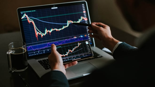 يدي شخص يمسك موبايل امام شاشة لابتوب فيها رسوم بيانية للتداول